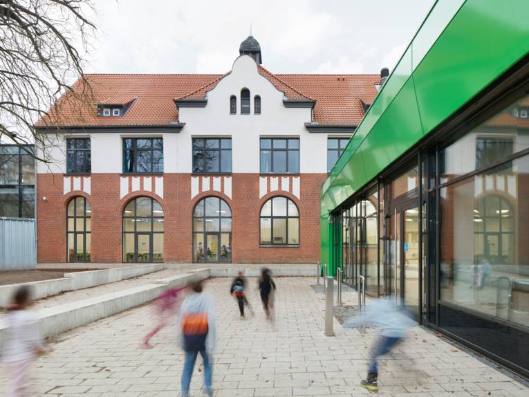 Rechts ein moderner flacher Bau mit gro&szlig;en Fenstern und einer gr&uuml;nen Fassade, hinten in der mitte ein altes Geb&auml;ude mit Schmuckelementen. Davor verwischt sind spielende Kinder zu erkennen.