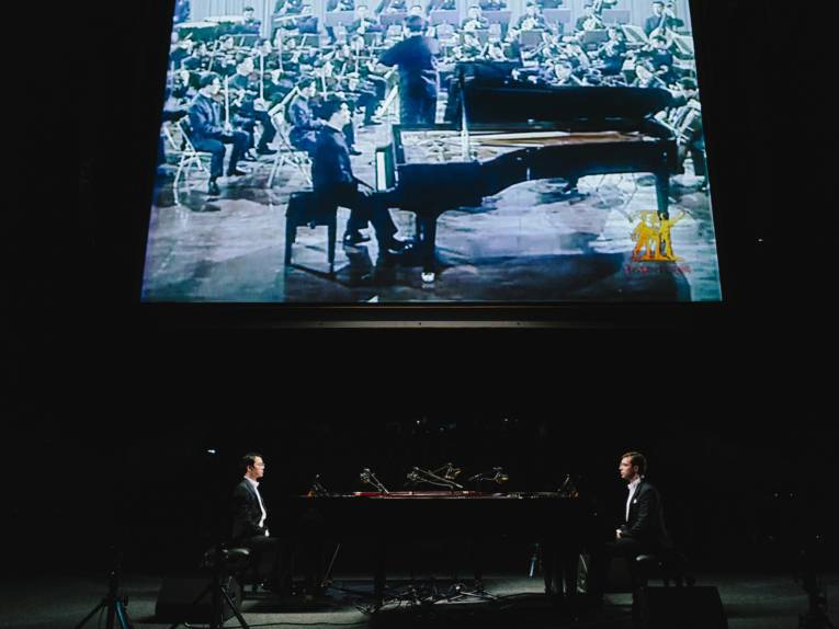 Zwei Pianisten mit Flügel auf einer Bühne, im Hintergrund ist auf einer Leinwand ein weiterer Pianist mit Flügel zu sehen.