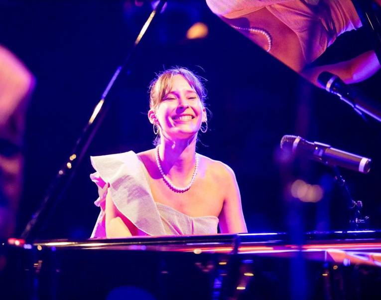 Frau in schulterfreiem Kleid mit hochgestecktem Haar, Ohrringen und Perlenkette sitzt an einem Konzertflügel.