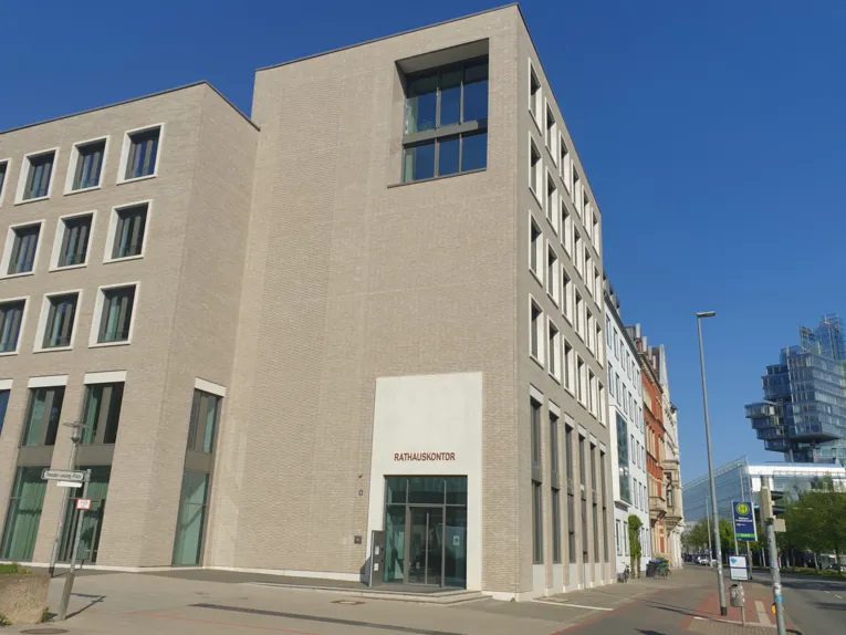 Auf dem Bild ist ein helles Gebäude mit einem roten Schriftzug "Rathauskontor" an einer Hauptstraße zu sehen. Im Hintergrund ist ein gläsernes Gebäude zu sehen bei wolkenlosen Himmel.