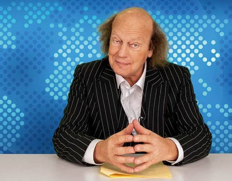 Mann mit langem seitlichen Resthaar in Hemd und Anzug sitzt mit aneinandergelegten Fingerspitzen an einem Tisch mit einem Manuskript.