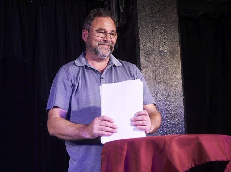 Lächelnder Mann mit kurzem Haar, Bart und Brille steht mit einem Papierstapel in den Händen an einem Tisch.