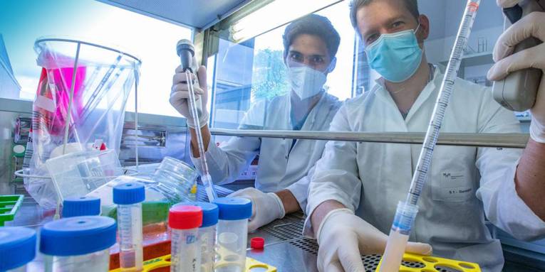 Zwei Männer in Arztkleidung mit Mundschutz und Gummihandschuhen hantieren mit Pipetten und Glaskolben.