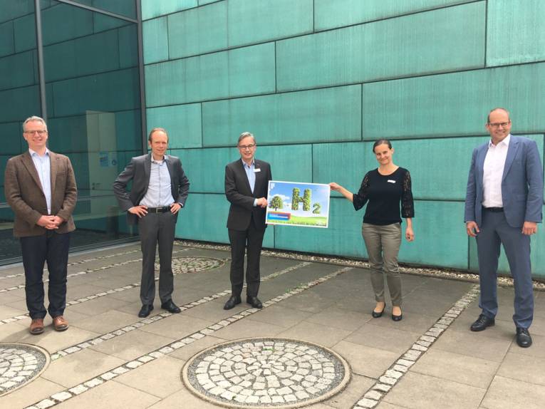 Fünf Personen stehen vor einem Gebäude, zwei davon halten gemeinsam ein Plakat auf dem "H2" steht.