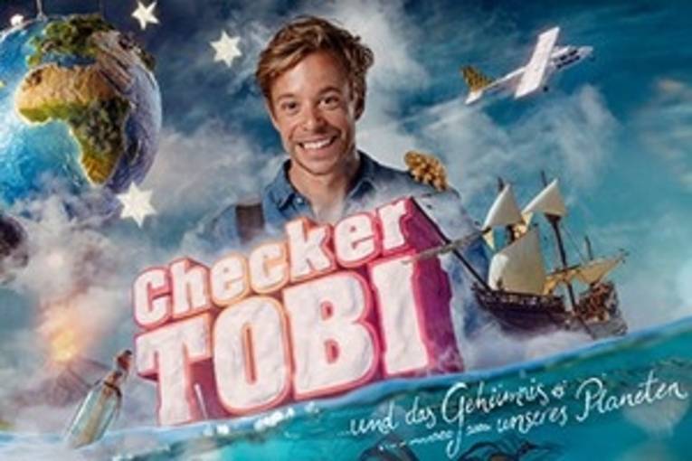 Checker Toby steht lächelnd hinter den Titel des Filmes, welcher auf dem Meer steht.