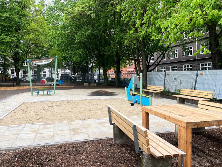 Ein Platz, vorn rechts eine Sitzecke aus Holz, in der Mitte ein großer in Bodenplatten eingelassener Sandkasten, rechts hinten ein buntes Spielgerät.