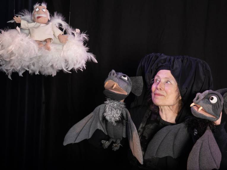 Dunkel gekleidete Puppenspielerin mit zwei Fledermaus-Handpuppen und einer Puppe, die eine älteren Mann als Engel auf einer Wolke darstellt.