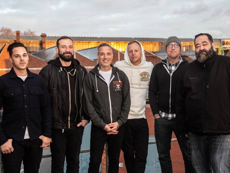 Sechs Männer stehen auf einem Flachdach. Im Hintergrund sind die Dächer von Industriegebäuden zu sehen. Alle schauen lächelnd in die Kamera.