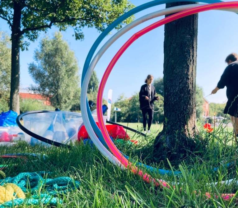 Hula Hoop reifen stehen an einen Baum gelehnt, daneben liegen Keulen und andere Zirkusutensilien. Im Hintergrund spielen Menschen mit einem Diabolo.