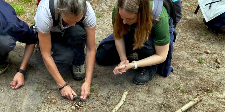 Zwei Frauen versuchen Feuer zu machen mit kleinen Ästen und Feuerstahl.
