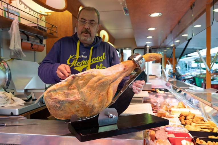 Ein Mann an einem Fleischwaren-Marktstand schneidet frischen Schinken von einem Schweinebein.