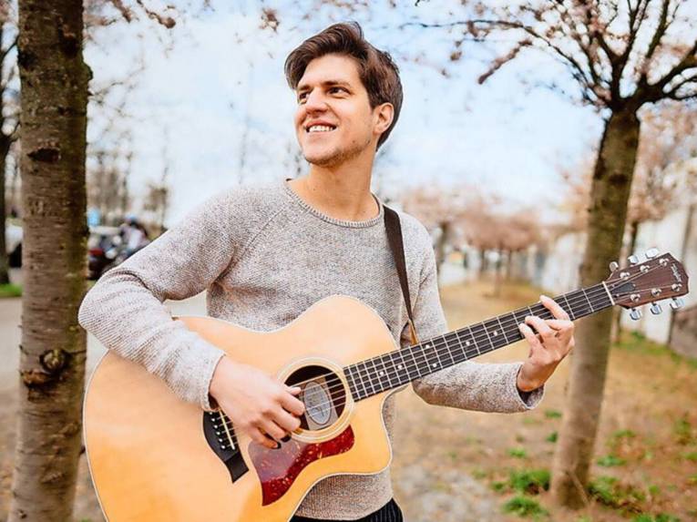 Lächelnder junger Mann spielt Gitarre, im Hintergrund Bäume und eine Straße