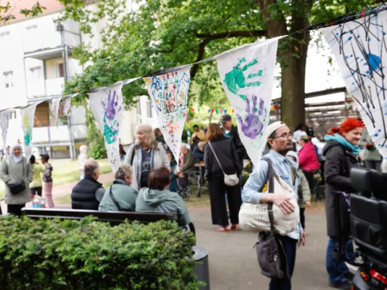 Menschen stehen und sitzen in einem Innenhof, &uuml;ber ihnen eine Girlande aus bunt bemalten F&auml;hnchen und Abdr&uuml;cken von Kinderh&auml;nden.