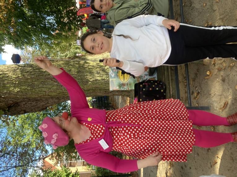 Clownin mit pinker Kleidung und roter Clownsnase h&auml;lt einen Stab auf dem etwas balanciert wird von einem Kind das in die Kamera blickt.