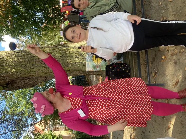 Clownin mit pinker Kleidung und roter Clownsnase h&auml;lt einen Stab auf dem etwas balanciert wird von einem Kind das in die Kamera blickt.