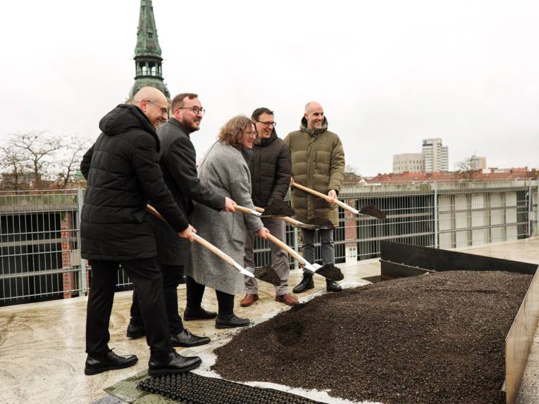 Fünf Personen stehen auf einem Dach vor einem Beet und halten Spaten mit Erde vor sich, hinter ihnen sind Dächer und ein Kirchturm zu sehen.