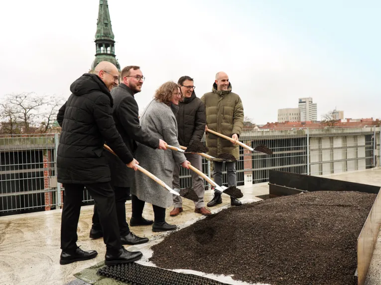 Fünf Personen stehen auf einem Dach vor einem Beet und halten Spaten mit Erde vor sich, hinter ihnen sind Dächer und ein Kirchturm zu sehen.