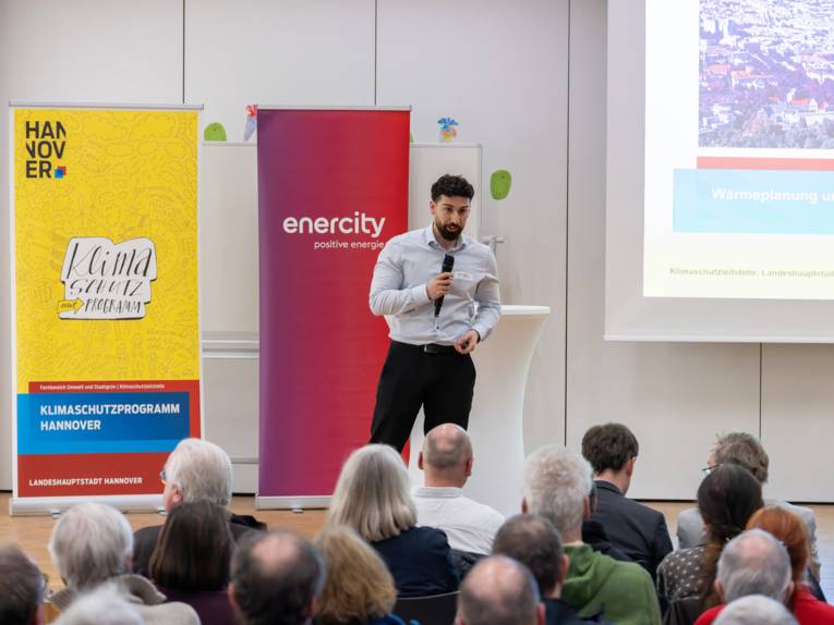 Ein Mann steht vor Publikum auf Stühlen auf einem Podest und spricht in ein Mikrofon. Hinter ihm stehen Roll-ups mit den Aufschriften Klimaschutzprogramm und enercity. An der Wand ist eine Präsentation mit dem Begriff Wärmeplanung zu sehen.