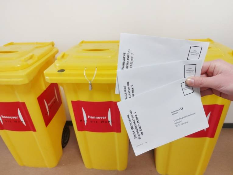 Eine Hand hält drei Briefumschläge, dahinter stehen drei gelbe Container oben mit offenen Einwurfschlitzen, vorn ein roter Aufkleber mit dem Logo Landeshauptstadt Hannover