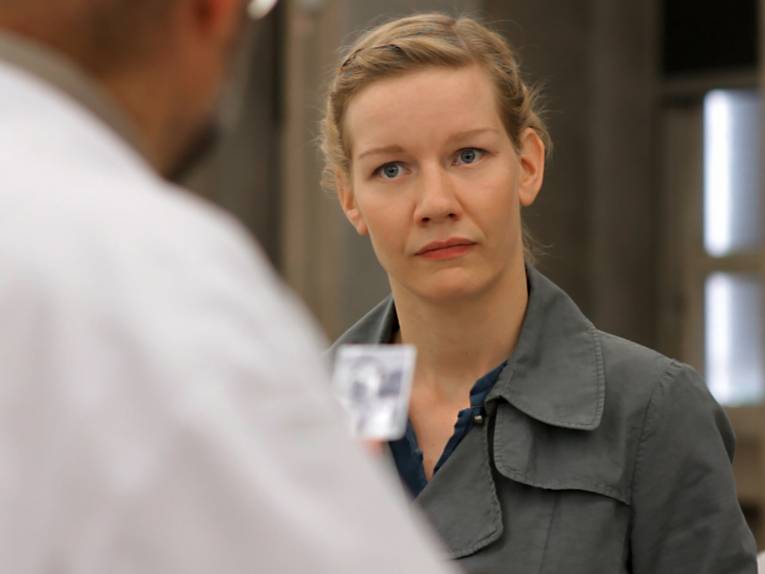 Das Bild zeigt eine Person in einem Innenraum in Nahaufnahme. Im Vordergrund ist unscharf eine weitere Person zu erkennen, während die fokussierte Person einen hellgrauen Mantel über dunkler Kleidung trägt. Der Hintergrund besteht aus warm beleuchteten architektonischen Elementen und großen Fenstern. Die geringe Schärfentiefe lenkt die Aufmerksamkeit auf die zentrale Figur und erzeugt eine ruhige, filmische Bildwirkung.