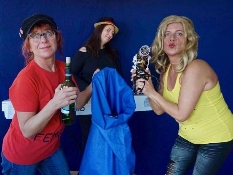 Drei Frauen, eine mit Weinflasche und Regencape in den Händen, eine an einem Keyboard, eine mit Föhn.