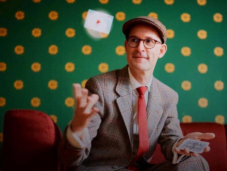 Mann mit Brille und Schiebermütze in Hemd, Schlips und Sakko wirft ein Herz-Ass in die Luft.