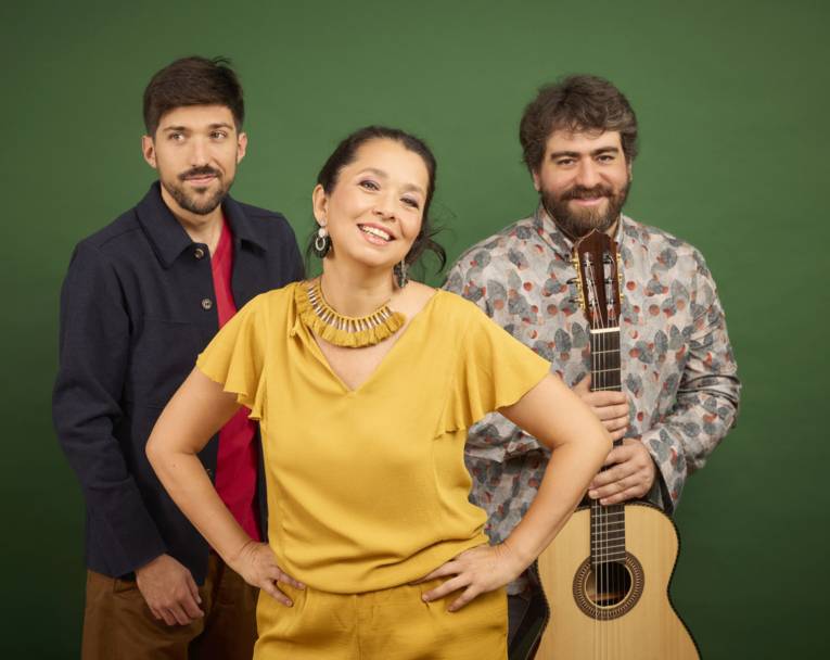 Vorn eine lächelnde Frau mit in die Hüften gestemmten Händen, hinter ihr zwei Männer, einer mit Gitarre