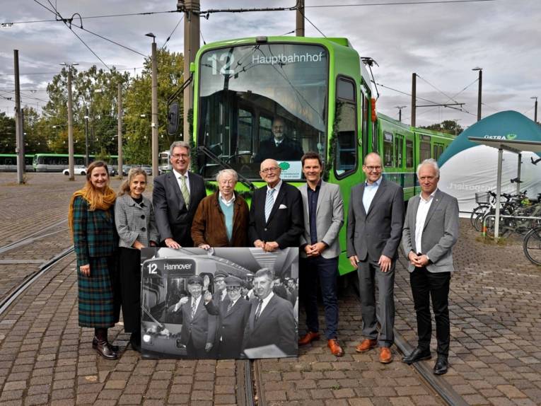 Eine Gruppe von Menschen steht vor einer Stadtbahn und präsentieren ein historisches Foto, auf dem die erste Stadtbahnfahrt von 1975 gezeit ist.