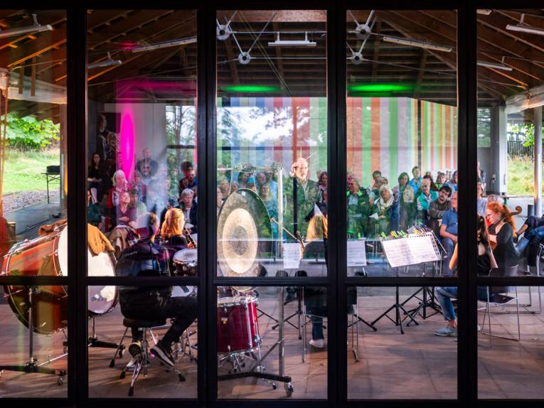 Blick von außen durch Glas auf ein spielendes klassisches Ensemble und Publikum.