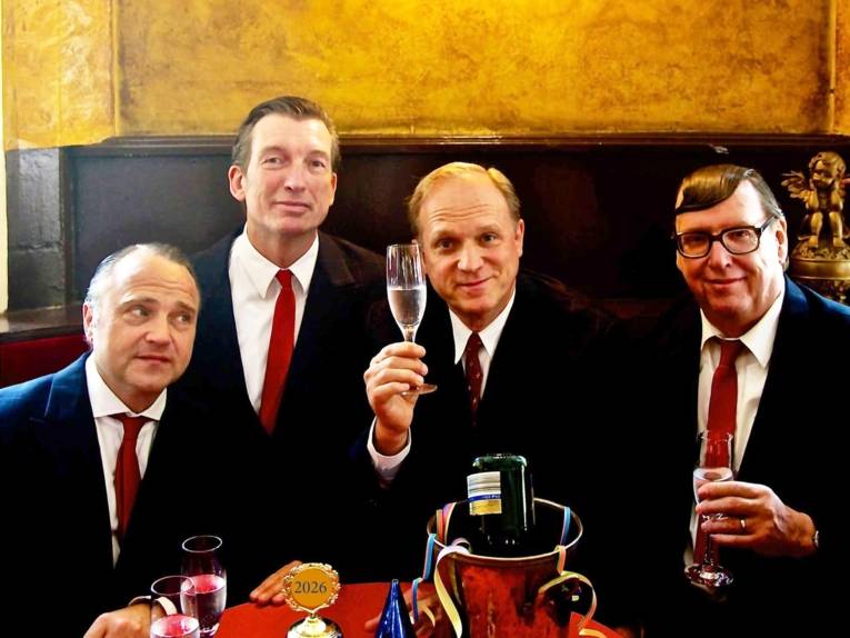 Zu sehen sind Ulrich Tukur und die Rhythmus Boys: Vier ältere Männer in schwarzen Anzügen und roten Krawatten, die in einem Restaurant zu sitzen scheinen und Sekt trinken.