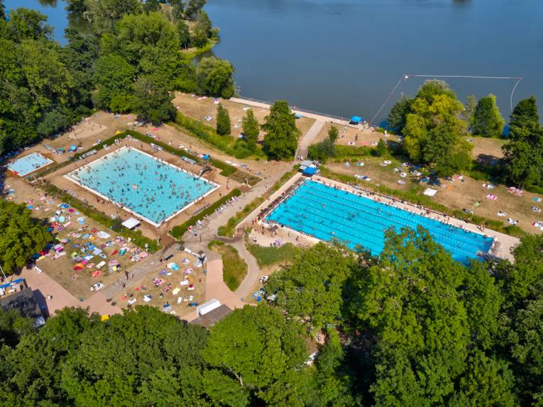 Luftbild eines gut besuchten Schwimmbades mit drei Becken, Liegewiesen und angrenzender Seefläche