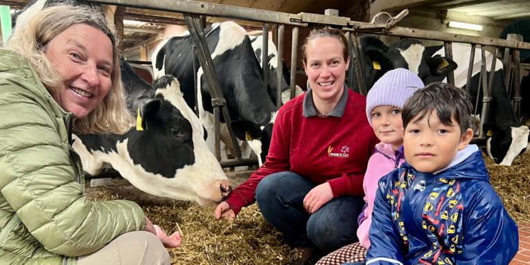 Zwei Frauen und zwei Kinder schauen in die Kamera, die vier Personen sind in einem Stall mit Kühen.