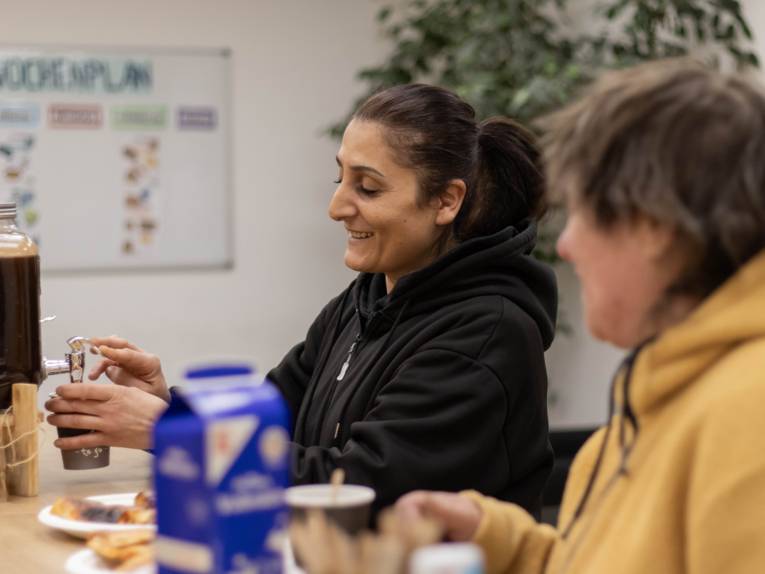 In einem Raum sitzen zwei Frauen an einem Tisch, eine schenkt Kaffee in einen Becher ein. Auf dem Tisch stehen Geschir und eine Milchtüte.