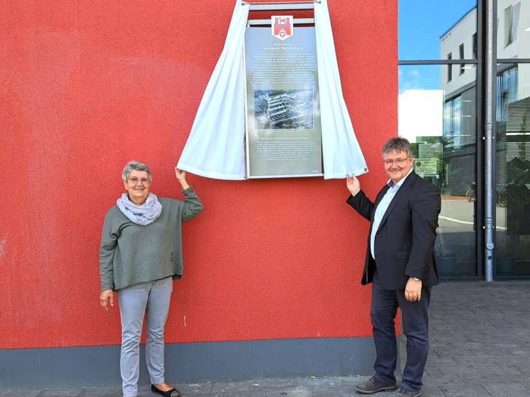 Brigitte Hurtzig, Vorsitzende der Arbeitsgemeinschaft Mühlenberg/Bornumer Vereine und Verbände e.V. (AMBV) und Bezirksbürgermeister Andreas Markurth enthüllen die neue Tafel am Stadtteilzentrum Weiße Rose.