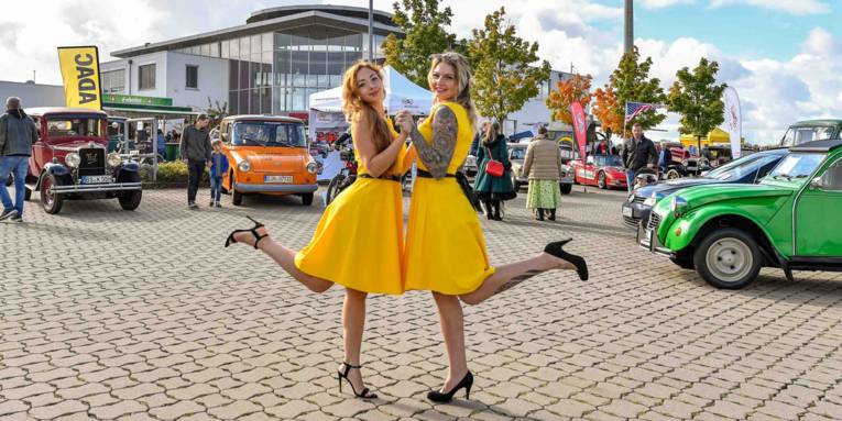 Zwei Frauen in gelben Kleidern posen vor Oldtimern.
