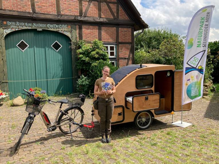 Eine Frau tr&auml;gt Outdoor-Kleidung, h&auml;lt Infomaterial in ihren H&auml;nden und steht vor einem besonderen Gespann aus E-Bike und Fahrradanh&auml;nger, einem kleinem Wohnmobil aus Holz.