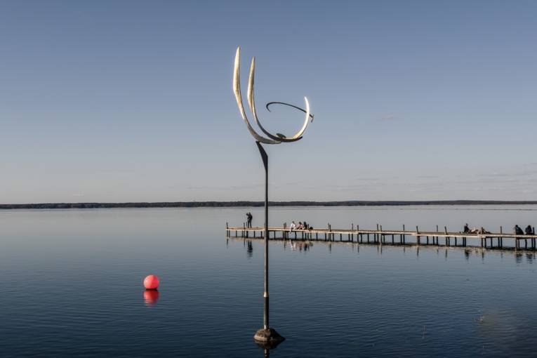 Wasser. Am Horizont ist Land. Von rechts kommt ein Steg ins Bild und im Vordergrund steht mittig eine Skulptur.