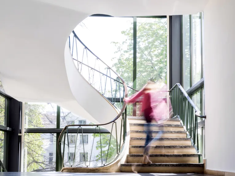 Eine Person steigt eine Treppe hinauf, das Treppenhaus ist hell und in weißer Farbe gestrichen, große Fenster geben den Blick nach draußen frei.