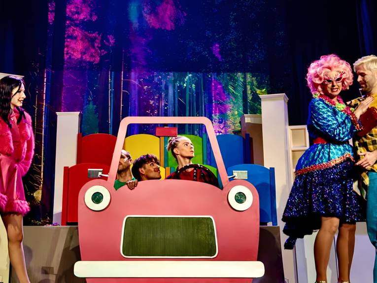 Bühnenszene: In der Mitte steht ein Pappauto, dahinter sind drei Personen zu sehen, links und rechts stehen weitere Personen