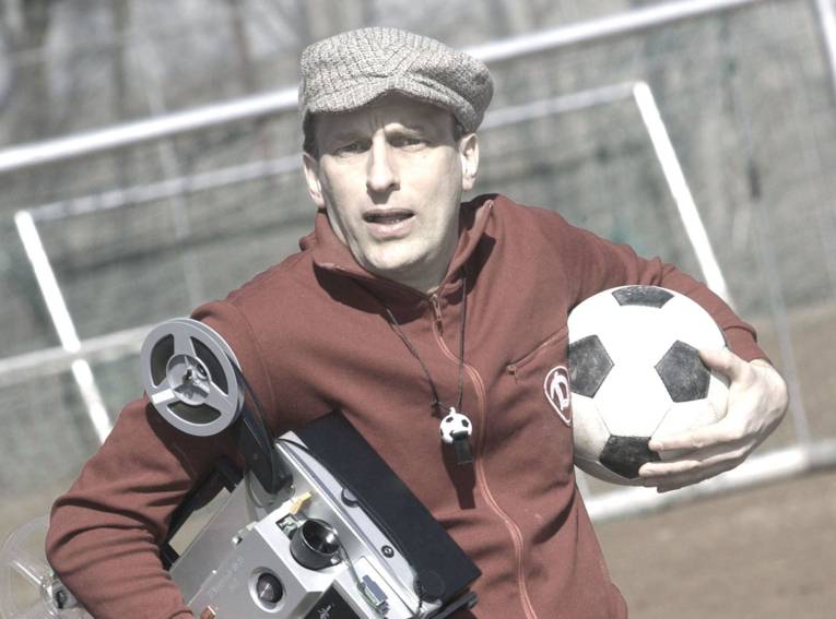 Mann mit Schiebermütze trägt einen Filmprojektor und einen Fußball unter den Armen.