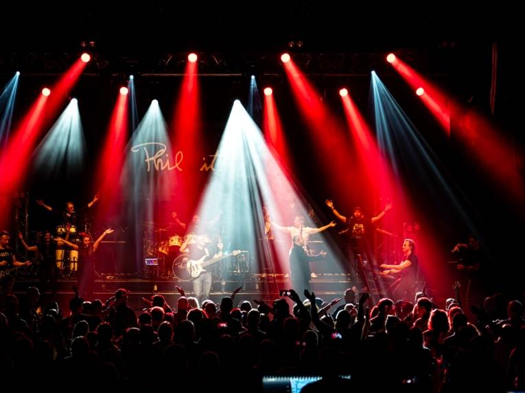 Zu sehen ist ein Konzert auf einer Bühne mit einer Gruppe Zuschauenden davor.