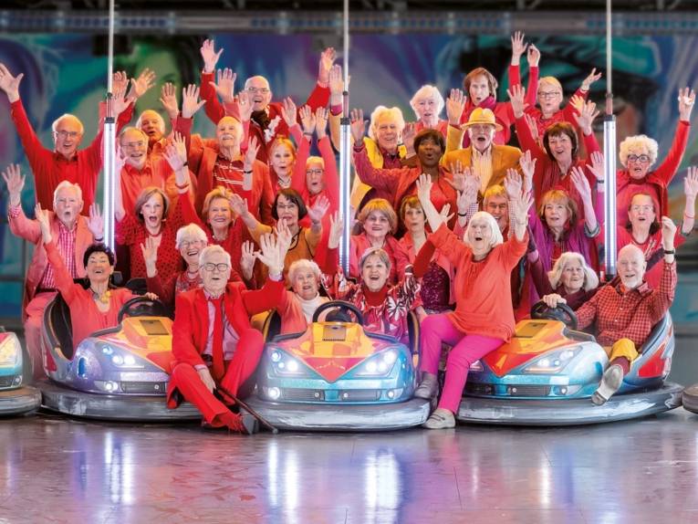 35 Sängerinnen und Sänger im Seniorenalter in roter Kleidung um drei Autoscooter verteilt.