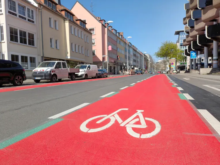 Eine Straße umringt von Gebäuden, vorn ein rot markierter langer Streifen mit einem weißen Fahrradsymbol, links parkende Autos.