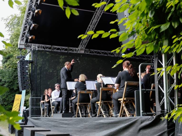 Musikerinnen und Musiker sind auf einer Bühne in einer grünen Kulisse. Der Dirigent leitet die Musizierenden an.