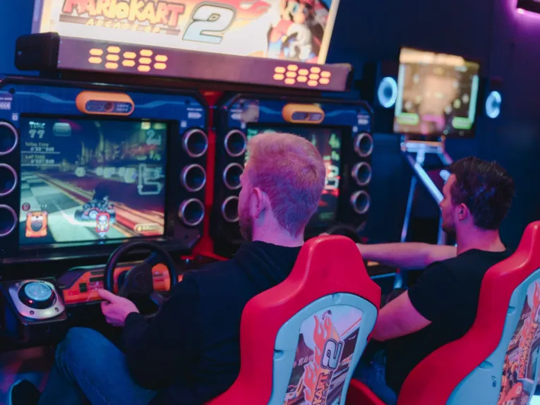 Zwei Männer sitzen in roten Arcade-Rennsitzen und spielen „Mario Kart Arcade GP 2“ in einer Spielhalle.