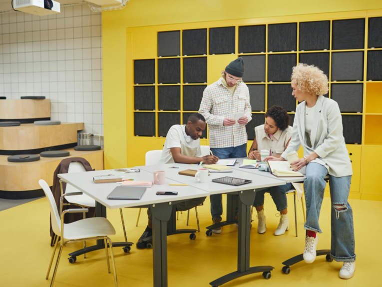 Vier Personen gruppieren sich um einen Gruppentisch. Anscheinend sind es zwei Frauen und zwei Männer. Sie tragen helle Kleidung. Sie konzentrieren sich auf Arbeitsunterlagen auf dem Tisch. Die Personen haben unterschiedliche Hautfarben. Der Raum, in dem sie sich befinden, ist in hellem gelb gehalten.