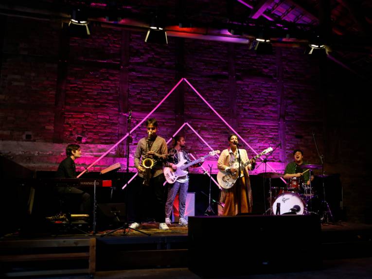 Vier Musiker und eine Musikerin geben auf einer Bühne ein Konzert. Im Hintergrund ist eine farbig angestrahlte Fachwerkmauer einer Scheune.