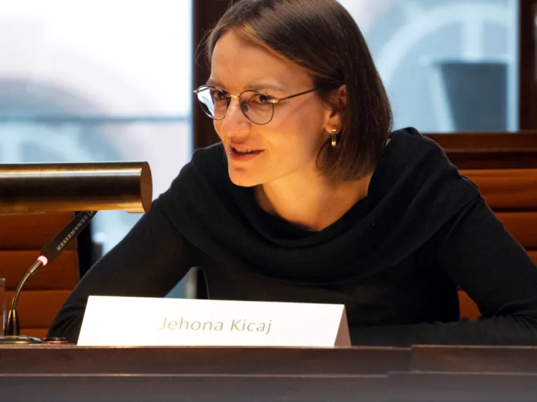 Eine Frau mit Brille und schwarzem Pullover sitzt leicht vorgebeugt an einem Tisch. Sie spricht in ein Tischmikrofon. Vor ihr ein Namensschild mit der Aufschrift Jehona Kicaj.