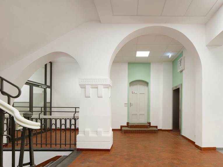 Links ein Treppenaufgang mit einem wei&szlig;en Gel&auml;nder in einen gro&szlig;en Raum mit zwei schmuckvollen B&ouml;gen, einer gro&szlig;en Glast&uuml;r und einer wei&szlig;en Zimmert&uuml;r.