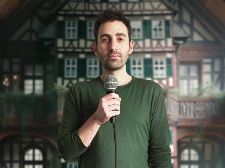 Zu sehen ist der Comedian Fabi Rommel in einem grünen Shirt vor einem Bild eines Fachwerkhauses. Er hält ein Mikrofon in der Hand.